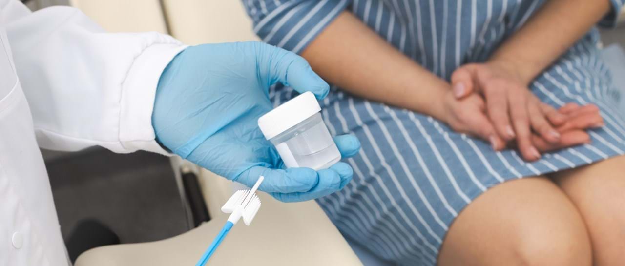 Helsepersonell med blå hansker holder en celleprøvebørste og en beholder for prøvetaking, mens en pasient sitter i bakgrunnen. Bildet illustrerer HPV-screening og livmorhalsprøvetaking.